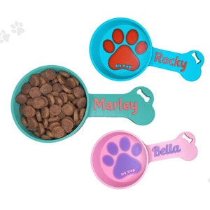 Two custom dog food scoops for measuring kibble. The blue scoop, personalized with the names Rocky holds 2/3 cup, while the pink scoop, labeled 'Bella,' holds 1/4 cup. Perfect for pet owners looking for personalized pet feeding accessories.