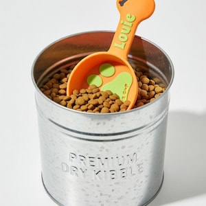 May include: A galvanized metal pet food container filled with dry kibble, with an orange scoop that has the name "Louie" in green. The scoop has a bone-shaped handle and a paw print design. The container is labeled "Premium Dry Kibble."