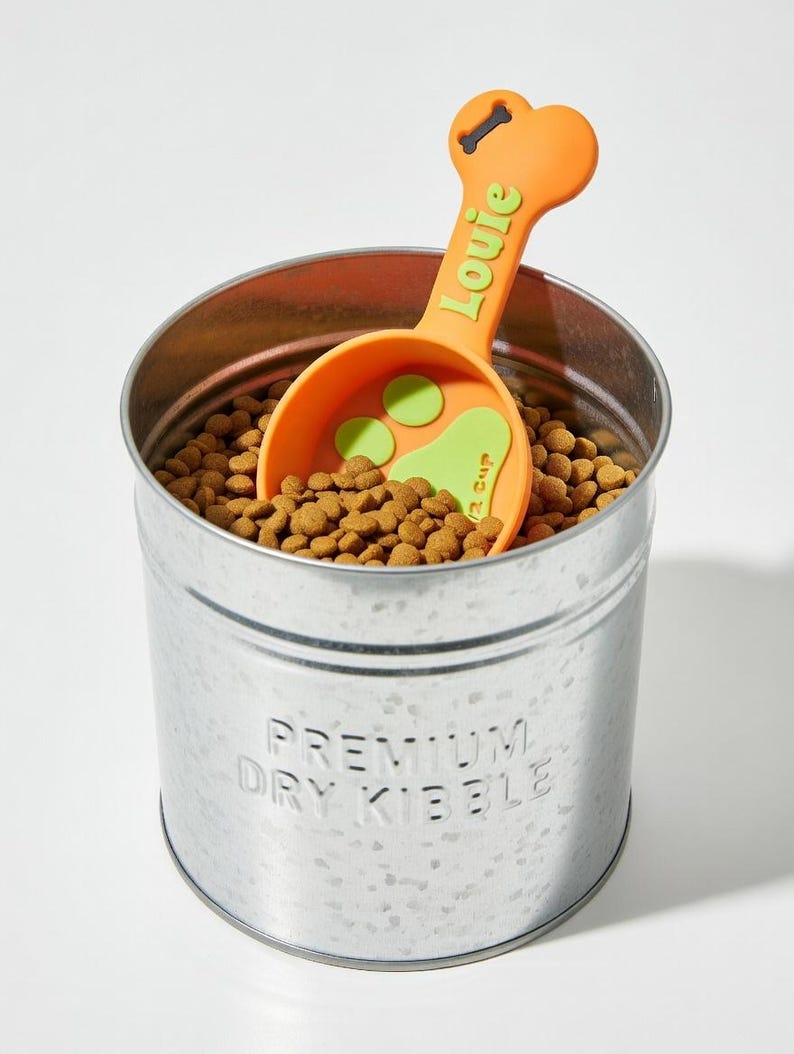 May include: A galvanized metal pet food container filled with dry kibble, with an orange scoop that has the name "Louie" in green. The scoop has a bone-shaped handle and a paw print design. The container is labeled "Premium Dry Kibble."