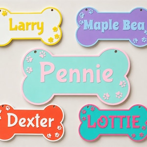 May include: Five colorful dog bone-shaped name tags. Each tag features a different name: "Larry", "Maple Bea", "Pennie", "Dexter", and "Lottie". The tags are in various colors, including yellow, purple, teal, orange, and pink, with paw print accents.