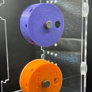 May include: Two cylindrical, magnetic, 3D-printed objects. The top one is purple with a black honeycomb edge. The bottom one is orange and has the text "PawtrN" and "Push to open". Both have a silver magnet.