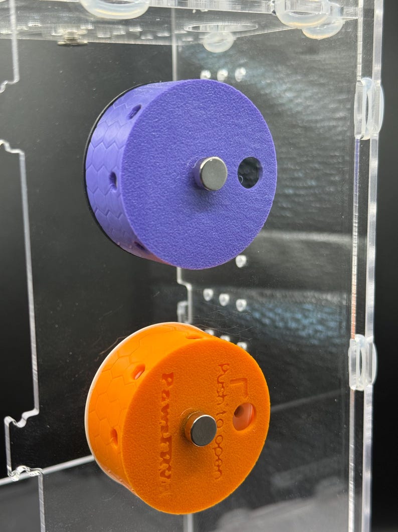 May include: Two cylindrical, magnetic, 3D-printed objects. The top one is purple with a black honeycomb edge. The bottom one is orange and has the text "PawtrN" and "Push to open". Both have a silver magnet.