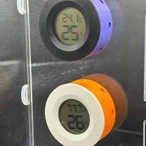 May include: Two digital hygrometers with round faces. One has a black bezel and purple honeycomb-patterned side, displaying 24.1&deg;C and 25%. The other has a white bezel and orange side, showing 77.7&deg;F and 26%.