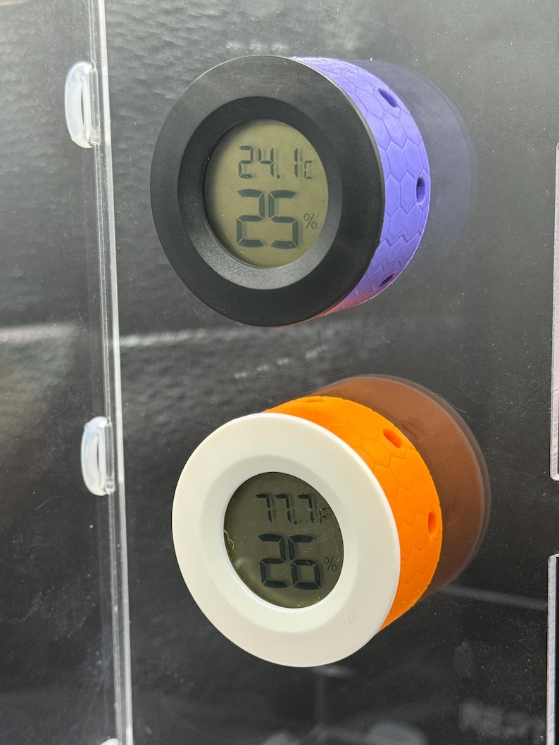 May include: Two digital hygrometers with round faces. One has a black bezel and purple honeycomb-patterned side, displaying 24.1&deg;C and 25%. The other has a white bezel and orange side, showing 77.7&deg;F and 26%.