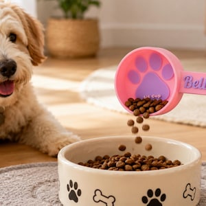 May include: A pink dog food scoop with a paw print design and the name "Bella" is pouring food into a beige bowl. The bowl has paw print and bone designs. A golden doodle dog is in the background.