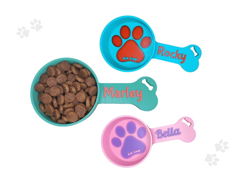 Two custom dog food scoops for measuring kibble. The blue scoop, personalized with the names Rocky holds 2/3 cup, while the pink scoop, labeled 'Bella,' holds 1/4 cup. Perfect for pet owners looking for personalized pet feeding accessories.