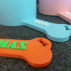 May include: Close-up of colorful dog bowl scoops. The scoops are in orange, light blue, and pink, each with a bone-shaped handle. The orange scoop has the name "MAX" in green letters. The other scoops have white lettering. These are likely pet supplies.