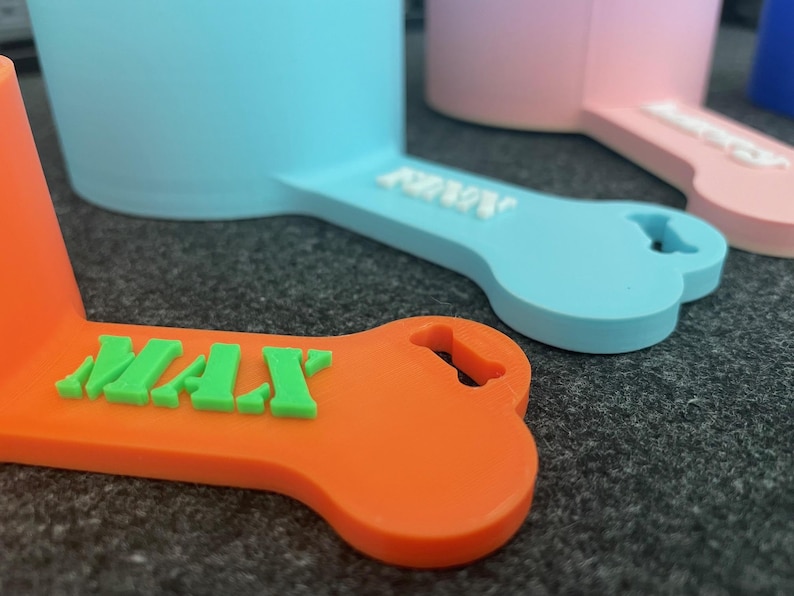 May include: Close-up of colorful dog bowl scoops. The scoops are in orange, light blue, and pink, each with a bone-shaped handle. The orange scoop has the name "MAX" in green letters. The other scoops have white lettering. These are likely pet supplies.