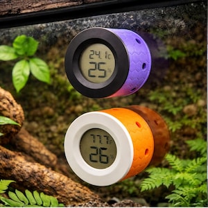May include: Two digital terrarium thermometers. One is purple and black, displaying 24.1°C and 25%. The other is orange and white, showing 77.7°F and 26%. Both have a textured, cylindrical design with a digital display.