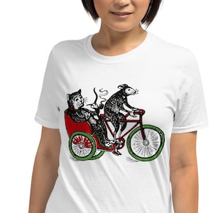 Può includere: Una maglietta bianca con un gatto nero e un topo che vanno in bicicletta. Il gatto indossa un cappello e fuma una pipa. Il topo indossa un cappello e tiene il manubrio. La bicicletta è rossa con ruote verdi.