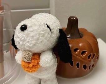 May include: A white and black crochet Snoopy doll holding an orange crochet pumpkin. A ceramic orange pumpkin with a brown stem is behind the doll. The Snoopy doll has black eyes and ears. The pumpkin has carved-out details.