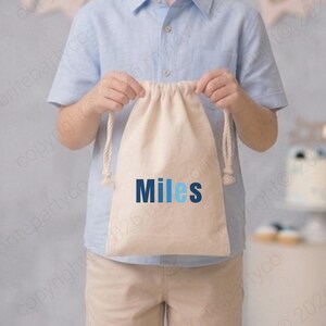 May include: A young boy smiles, holding a beige drawstring bag with the name "Miles" in blue. He wears a light blue button-down shirt and khaki shorts, standing in front of a backdrop of blue, white, and black balloons. A cake and banner that says "DAY" are in the background.