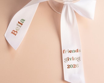 May include: A clear glass coupe with a white satin ribbon tied around the stem. The ribbon features the name Bella printed in orange and green on one tail, and the text friends giving 2026 printed in orange and green on the other tail.