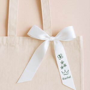 May include: A beige canvas tote bag featuring a white satin ribbon tied in a bow. The ribbon is printed with dark green line art of a butterfly, two small flowers, a crown, and the name Rachel in a simple sans-serif font on the right tail.