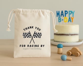 May include: A cream-colored drawstring bag with the text "THANK YOU FOR RACING BY Wyatt's First Birthday" and crossed checkered flags. A cake with a "HAPPY B-DAY" topper and wooden toys are in the background.
