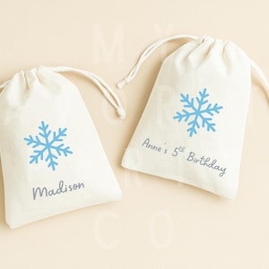 May include: Two off-white drawstring bags with light blue snowflake designs. One bag has the name "Madison" printed on it, and the other says "Anne's 5th Birthday". The bags are on a light beige background.