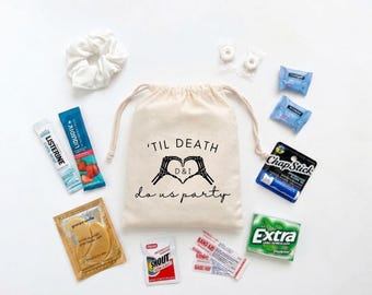 May include: A beige drawstring canvas bag printed with the text 'TIL DEATH do us party' and a skeleton hand heart graphic with 'D & I' inside. Surrounding the bag are a white scrunchie, Listerine, Liquid IV, ChapStick, Extra gum, gold eye masks, Shout wipes, Band-Aids, and mints.