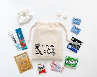 May include: A beige drawstring canvas bag printed with a skeleton hand holding a martini glass and the text '2027 Til Death do us Party Tiffany's Bachelorette'. Surrounding the bag are a white scrunchie, Listerine, Liquid IV, ChapStick, Extra gum, gold eye patches, Shout wipes, Band-Aids, and two mints.