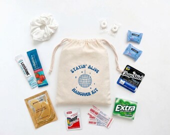 May include: A beige drawstring canvas bag printed with a blue disco ball and the text Stayin' Alive Hangover Kit, surrounded by a white scrunchie, Listerine, Liquid IV, ChapStick, Extra gum, gold eye masks, Shout wipes, Band-Aids, and mints.