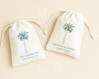 May include: Two off-white fabric drawstring pouches featuring palm tree illustrations. The left pouch displays blue text reading THE ANDERSONS SAN DIEGO, CA. The right pouch features green text reading AMANDA & DANIEL August 1, 2026.