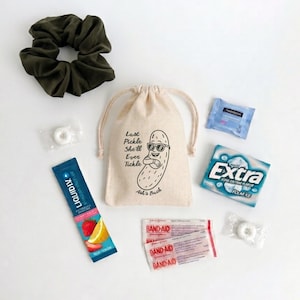 Puede incluir: Una bolsa beige con cordón que muestra un pepinillo de dibujos animados con gafas de sol y el texto Last Pickle She'll Ever Tickle Ash's Bach, un coletero verde oliva, un sobre de Liquid I.V., un paquete de chicles Extra, dos tiritas y dos caramelos de menta.