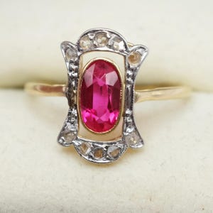 May include: A gold ring featuring a central oval-cut vibrant pink gemstone set in a yellow gold bezel. The stone is surrounded by a silver-toned frame embellished with small, round, clear stones, creating a rectangular, open-work design.