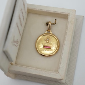 May include: A round gold pendant featuring a central cross with a small diamond, the French text QU'HIER and QUE DEMAIN, and three small rectangular red rubies set horizontally. The pendant is displayed inside a small, open, cream-colored box.