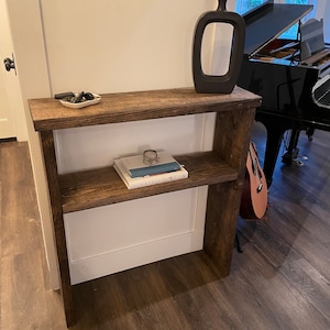 May include: A rustic wooden console table with two shelves. The table is stained a dark brown colour and has a distressed finish. The top shelf has a small white dish with some decorative stones. The bottom shelf has a stack of books and a guitar leaning against the table.