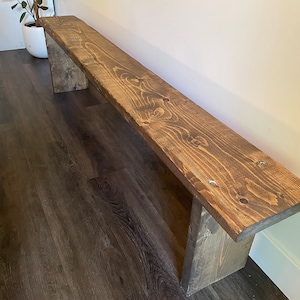 May include: A long, wooden bench with a dark brown stain. The bench has a simple design with two legs at each end. The bench is sitting on a dark brown wood floor.