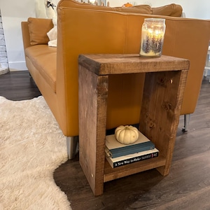 May include: A rustic, wooden side table with a lower shelf holding books and a small pumpkin. A lit candle in a glass jar sits on top. The table is next to a tan leather couch and a fluffy rug.