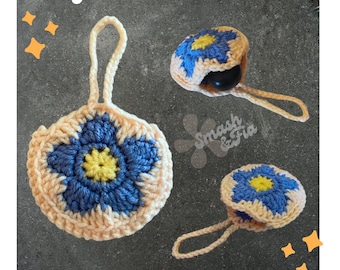 May include: Two crocheted flower-shaped pouches with a blue and yellow flower design. The pouches are made with a beige yarn and have a loop for hanging. The text "Easy No Sew Pattern" is at the top of the image. The text "Earbud Pouch - Accessory Bag" is at the bottom of the image.