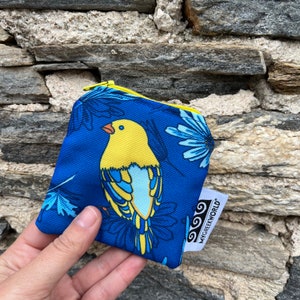 Op de afbeelding: Een kleine blauwe ritszak met een gele vogel en blauwe bloemen erop gedrukt. De tas heeft een gele rits en een wit label met de tekst "MY GREEK WORLD".