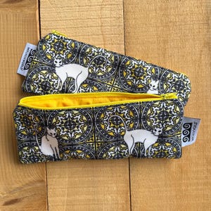 May include: A yellow and black zippered pouch with a white cat design. The pouch has a yellow lining and a zipper pull with a black and white swirl design. The pouch has the text "Wickerworld" on the side.