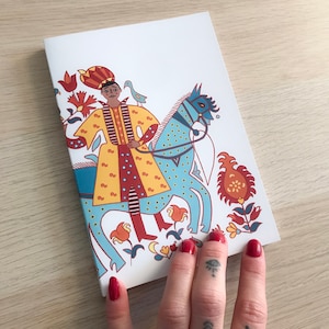 Può includere: Un quaderno bianco con un'illustrazione colorata di una persona a cavallo di un cavallo blu. La persona indossa una veste gialla e rossa con una corona in testa. L'illustrazione è circondata da fiori e foglie.