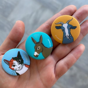 Könnte beinhalten: Drei runde Anstecknadeln mit Comic-Tierillustrationen. Der erste Knopf ist blau mit einer weißen Katze mit orangefarbenen und schwarzen Markierungen. Der zweite Knopf ist blau mit einem braunen Esel. Der dritte Knopf ist gelb mit einer blauen Ziege.