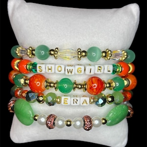 May include: Five beaded bracelets with gold accents and various colorful beads, including green, orange, and white letter beads spelling "SHOWGIRL" and "ERA". The bracelets are displayed on a white pillow against a black background.