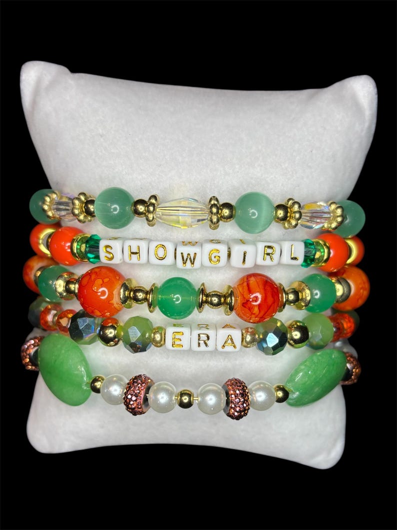 May include: Five beaded bracelets with gold accents and various colorful beads, including green, orange, and white letter beads spelling "SHOWGIRL" and "ERA". The bracelets are displayed on a white pillow against a black background.