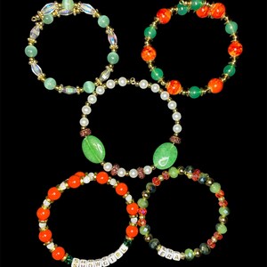 May include: Five beaded bracelets in various designs. One bracelet features the text "SHOWGIRL" and another reads "ERA". The bracelets include green, orange, and clear beads, with gold accents and a pearl bracelet.