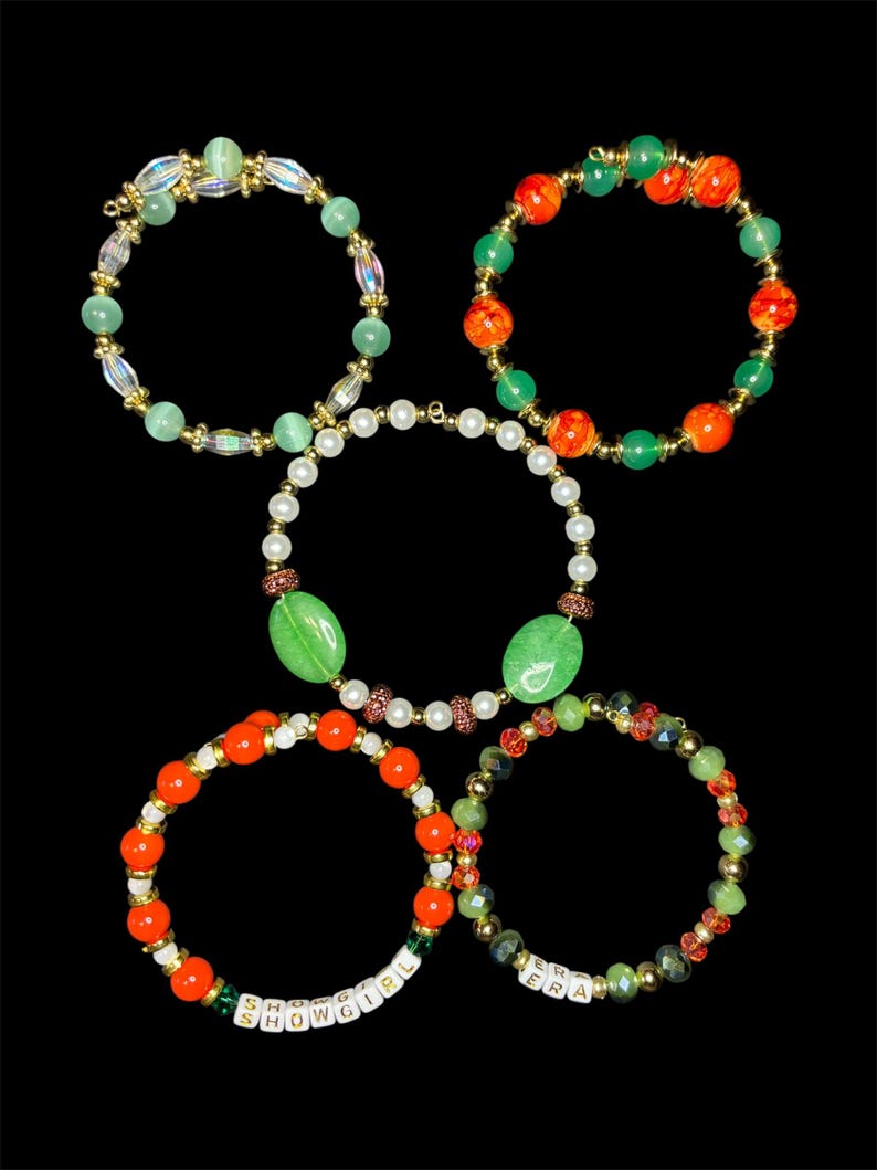 May include: Five beaded bracelets in various designs. One bracelet features the text "SHOWGIRL" and another reads "ERA". The bracelets include green, orange, and clear beads, with gold accents and a pearl bracelet.