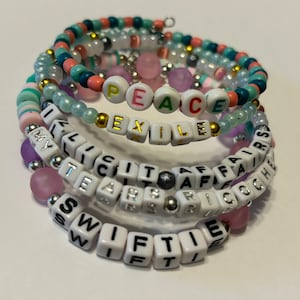 May include: A set of five colorful beaded bracelets with white letter beads spelling out the words "PEACE", "EXILED", "LICIT AFFAIRS", "TEARS OR", and "SWIFTIE".