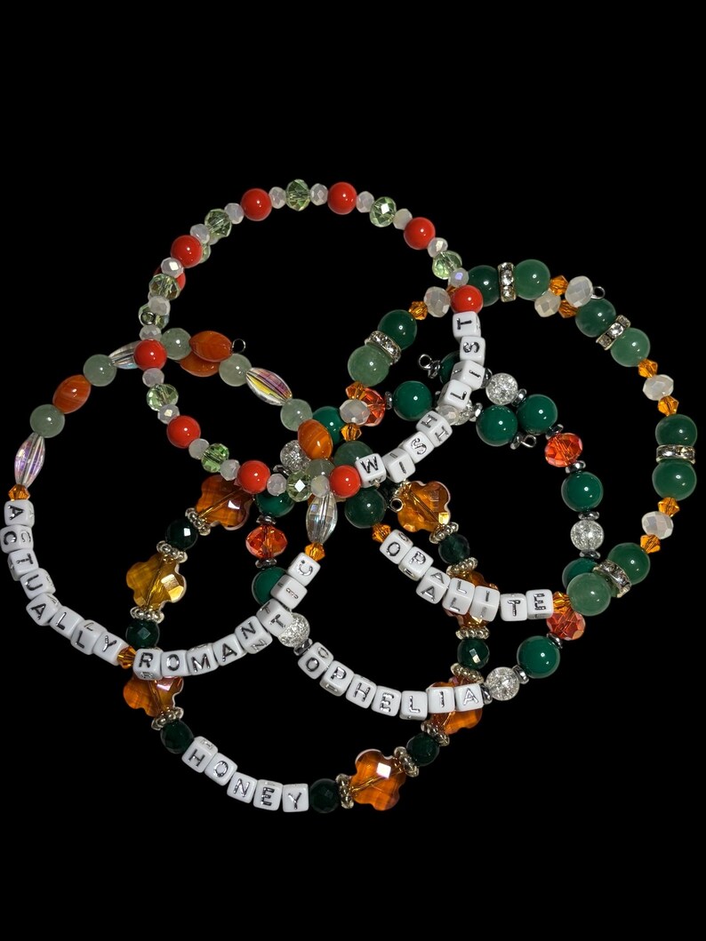 Pu&ograve; includere: Quattro braccialetti di perline su sfondo nero. I braccialetti presentano una variet&agrave; di perline colorate, tra cui rosse, verdi, arancioni e trasparenti. Alcuni braccialetti hanno perline con lettere bianche che compongono frasi come "ACTUALLY ROMANTIC" e "WISH OF OPHELIA".