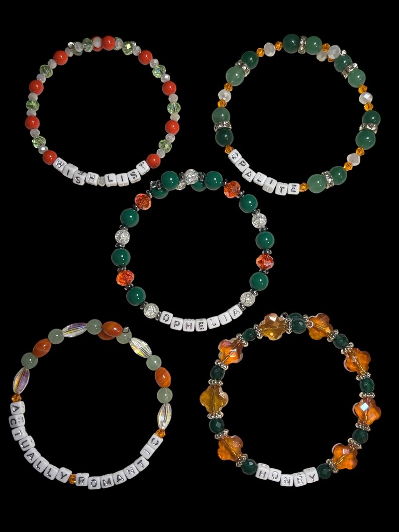 Pu&ograve; includere: Cinque braccialetti di perline con vari schemi di colori e testo. Un braccialetto dice "WISH LIST" con perline rosse e bianche. Un altro dice "OPALITE" con perline verdi. Il terzo dice "ACTUALLY ROMANTIC" con perline arancioni. Il quarto dice "HONEY" con perline arancioni.