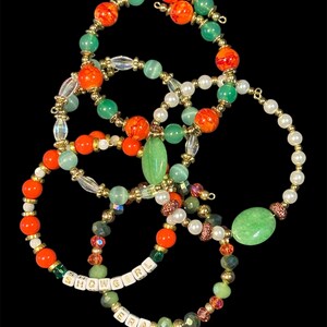 May include: A collection of beaded bracelets in various colors, including orange, green, white, and gold. Some bracelets feature the text "SHOWGIRL ERA" spelled out with white letter beads. The bracelets are arranged against a black background.