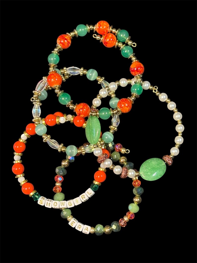 May include: A collection of beaded bracelets in various colors, including orange, green, white, and gold. Some bracelets feature the text "SHOWGIRL ERA" spelled out with white letter beads. The bracelets are arranged against a black background.