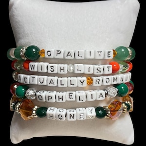 Pu&ograve; includere: Cinque braccialetti di perline con perline di lettere bianche che compongono "OPALITE", "WISH LIST", "ACTUALLY ROMA", "OPHELIA" e "HONEY". I braccialetti presentano perline verdi, arancioni e trasparenti e sono esposti su un cuscino bianco.