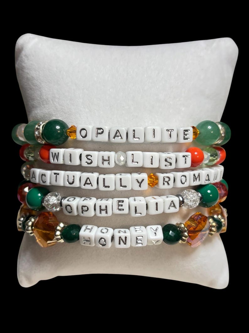 Pu&ograve; includere: Cinque braccialetti di perline con perline di lettere bianche che compongono "OPALITE", "WISH LIST", "ACTUALLY ROMA", "OPHELIA" e "HONEY". I braccialetti presentano perline verdi, arancioni e trasparenti e sono esposti su un cuscino bianco.