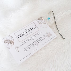 May include: A white card with gold lettering that reads "The Tesseract Silktie Atelier" and describes the Tesseract space stone. The card is on a white fluffy surface with a silver bookmark with a blue crystal charm.
