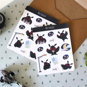 Pode incluir: Duas folhas de adesivos com ilustrações de Mothman. Cada folha tem um cabeçalho preto com a palavra "Mothman". Os adesivos retratam Mothman com um mapa, uma lanterna e um poste de luz. Os adesivos estão em um fundo azul claro.