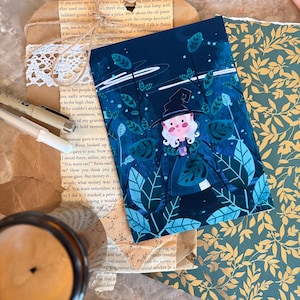 May include: A rectangular art print featuring a character with white hair and a black hat standing in a dark blue forest with teal leaves. The print is placed on brown paper with white lace trim, alongside a dark amber candle and gold leaf paper.