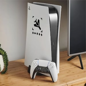 May include: A white PlayStation 5 console with a black design and a matching white controller. The console is upright on a wooden surface, with a black and silver trim. The controller is in front of the console.